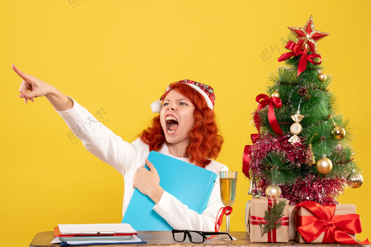 文件前视图：女医生坐在桌子后面 手里拿着文件 背景是黄色的圣诞树和礼盒背景人快乐