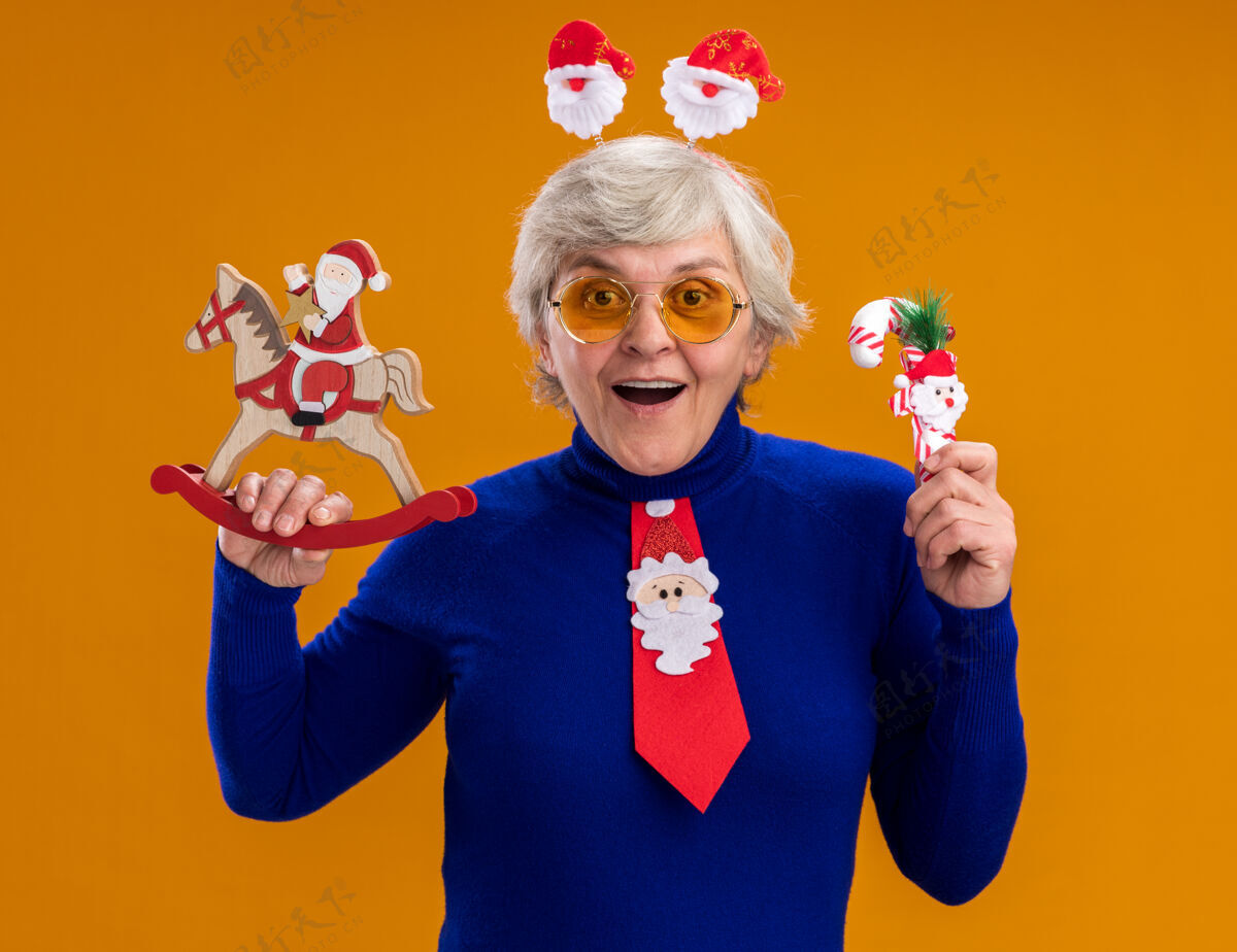惊讶戴着太阳眼镜 戴着圣诞老人头带 系着圣诞老人领带的惊讶的老妇人把圣诞老人抱在摇马装饰上 把糖果手杖孤立在橙色背景上 留有复制空间手杖老人圣诞老人