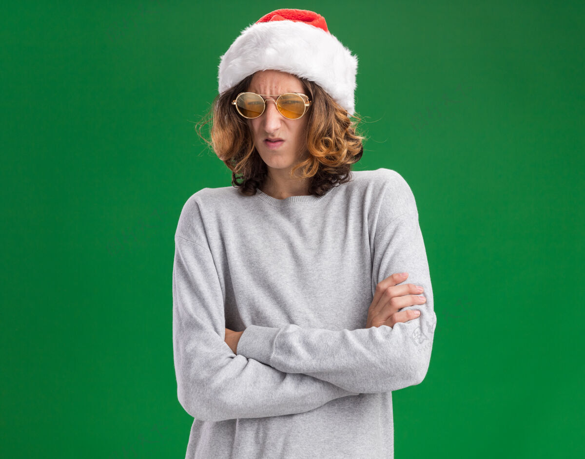 圣诞节不满的年轻人戴着圣诞老人帽 戴着黄色眼镜 皱着眉头 双臂交叉地站在绿色背景上 看着摄像机眼镜站着脸