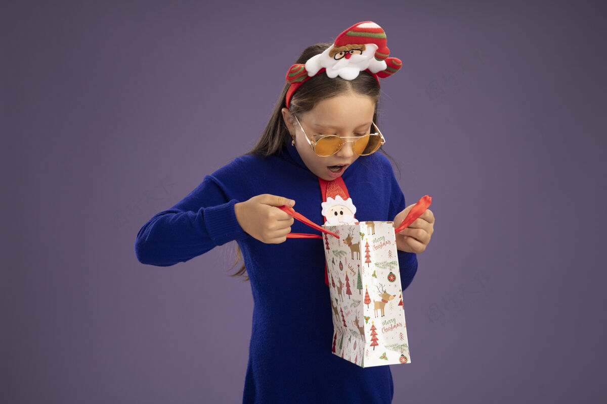 圣诞节小女孩穿着蓝色高领毛衣 系着红色领带 头上戴着有趣的圣诞镶边 手里拿着一个装有圣诞礼物的纸袋 站在紫色的背景下惊讶地看着里面拿着边缘里面