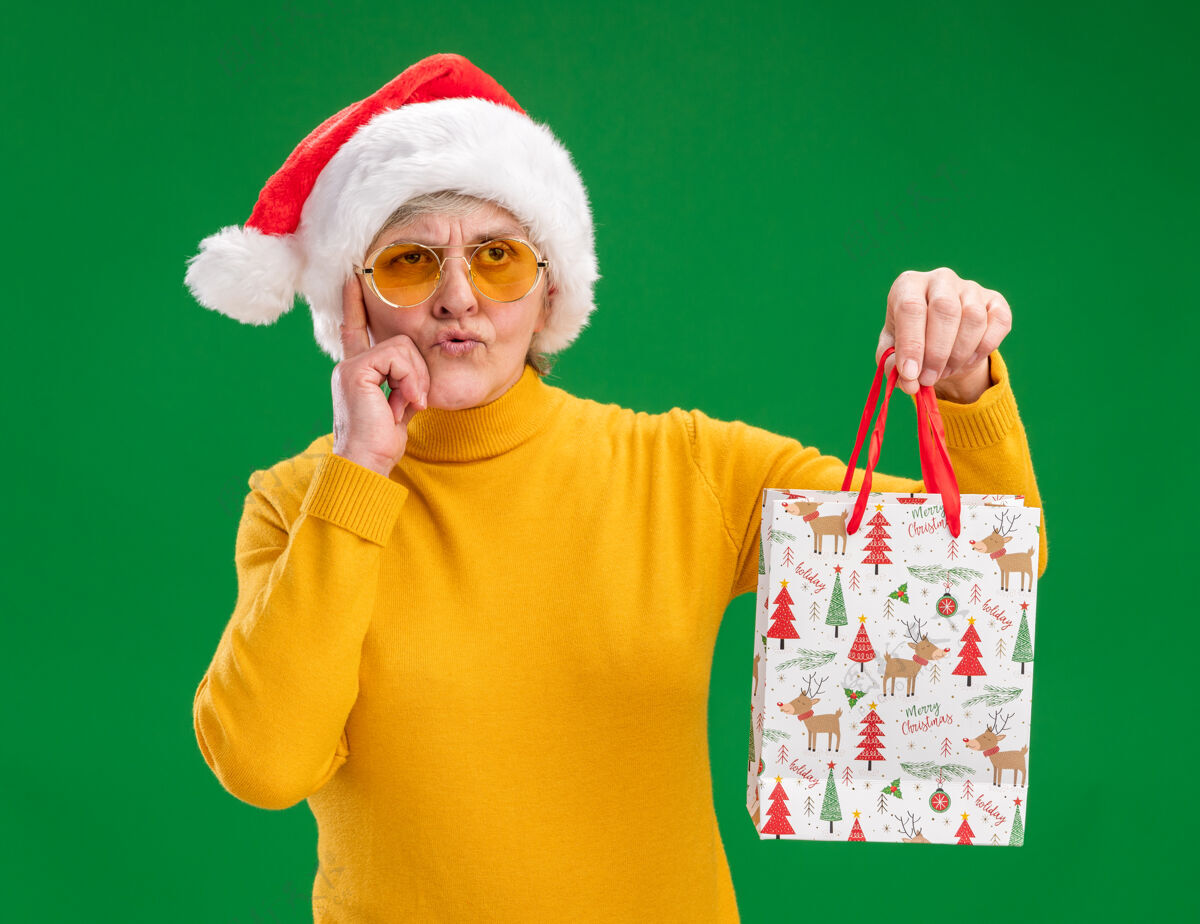 看戴着太阳眼镜 戴着圣诞帽 体贴的老妇人把手指放在太阳穴上 拿着纸礼包 看着隔离在绿色背景上的一面 还有复印空间侧圣诞节帽子