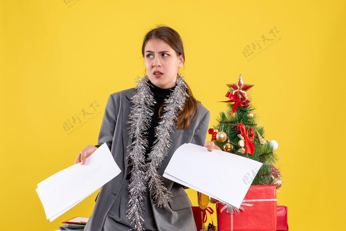 成人前视图年轻女孩拿着文件靠近圣诞树和礼物鸡尾酒年轻女孩树花束