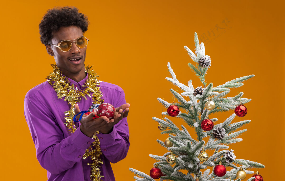 花环令人印象深刻的年轻非裔美国人戴着眼镜 脖子上戴着金箔花环 站在装饰过的圣诞树旁 拿着圣诞饰品 看着孤立在橙色背景上的圣诞饰品镀金圣诞树圣诞