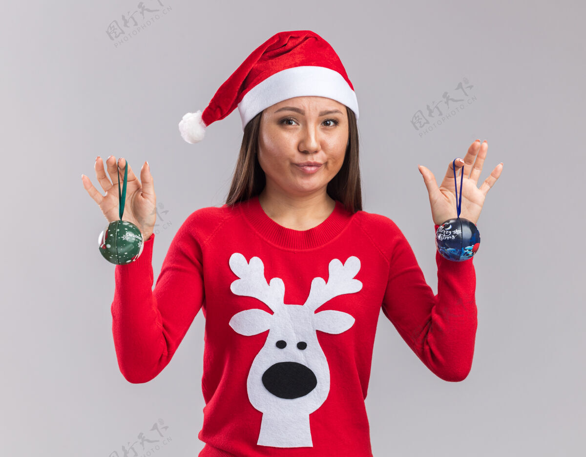 球高兴的年轻亚洲女孩戴着圣诞帽与毛衣举行圣诞树球隔离在白色背景树举行年轻