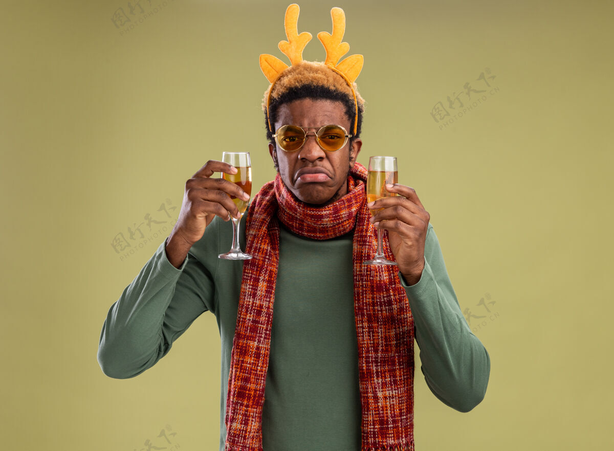 角一个非洲裔美国人 戴着滑稽的边 戴着鹿角 脖子上围着围巾 手里拿着两杯香槟 神情悲伤地看着摄像机 噘着嘴唇站在绿色的背景上站着边缘鹿