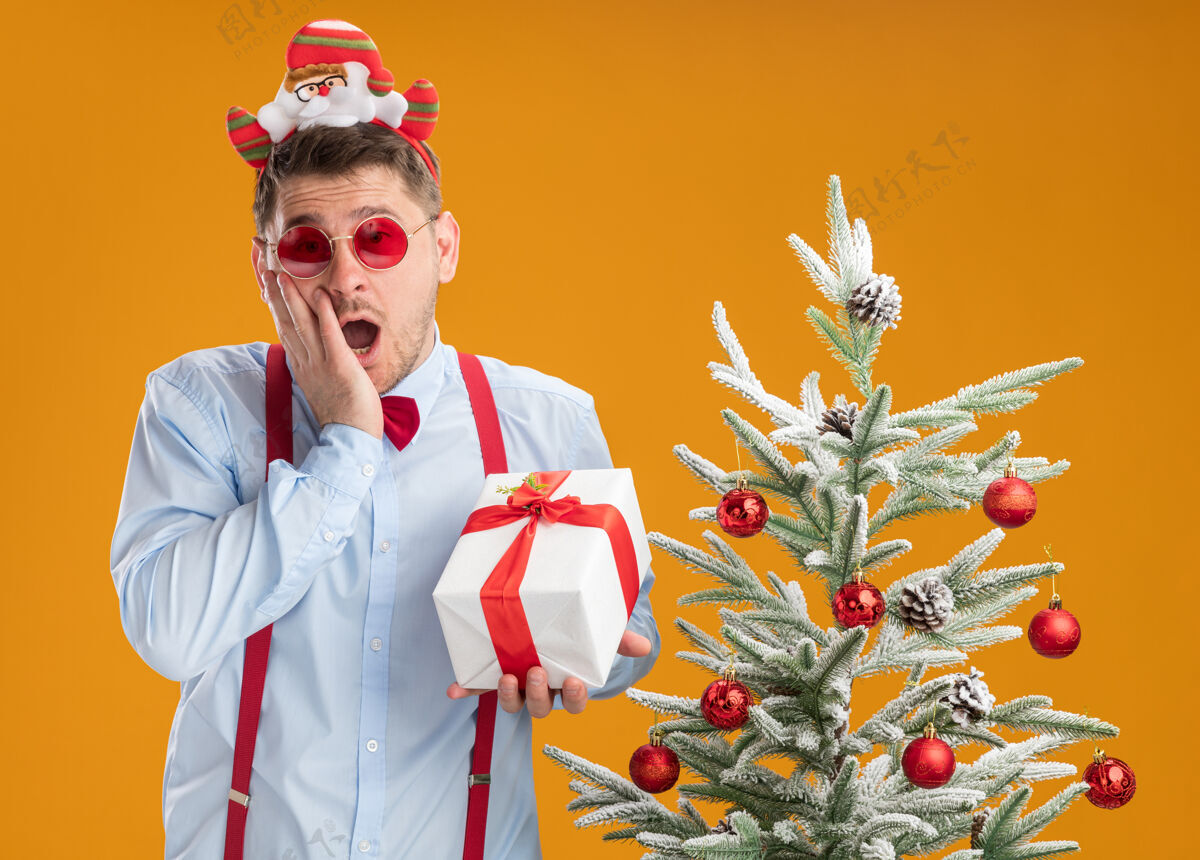 兴奋戴着吊带领结的年轻人站在圣诞树旁 戴着圣诞老人和红眼镜 手里拿着一份礼物 在橙色的背景下快乐而兴奋圣诞节眼镜领带