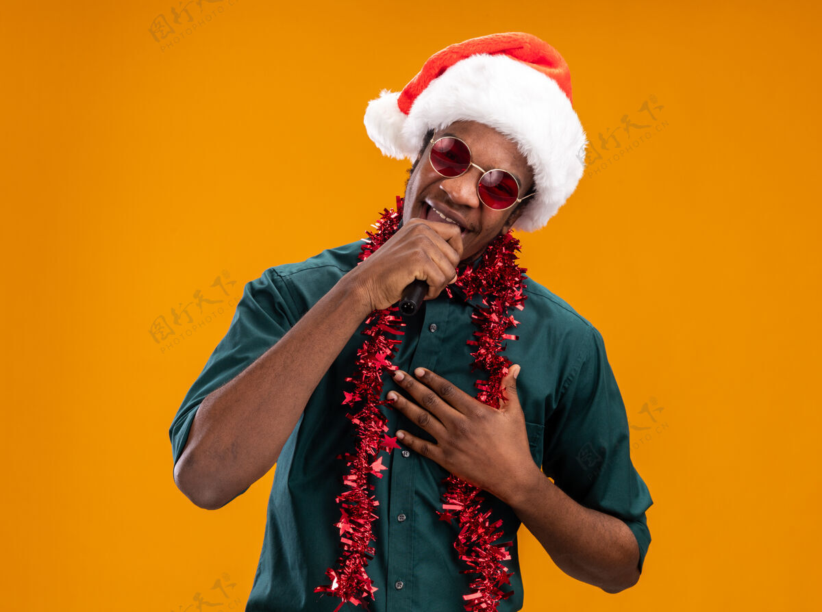圣诞老人戴着圣诞帽 戴着花环 戴着眼镜的非裔美国人站在橙色背景下 对着麦克风快乐地唱歌麦克风眼镜站着