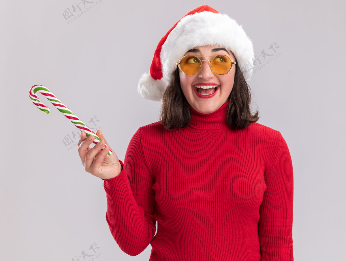 手杖穿着红色毛衣 戴着圣诞帽 戴着眼镜 手持糖果手杖 站在白色背景下 开心地看着一边的年轻女孩脸拿着毛衣