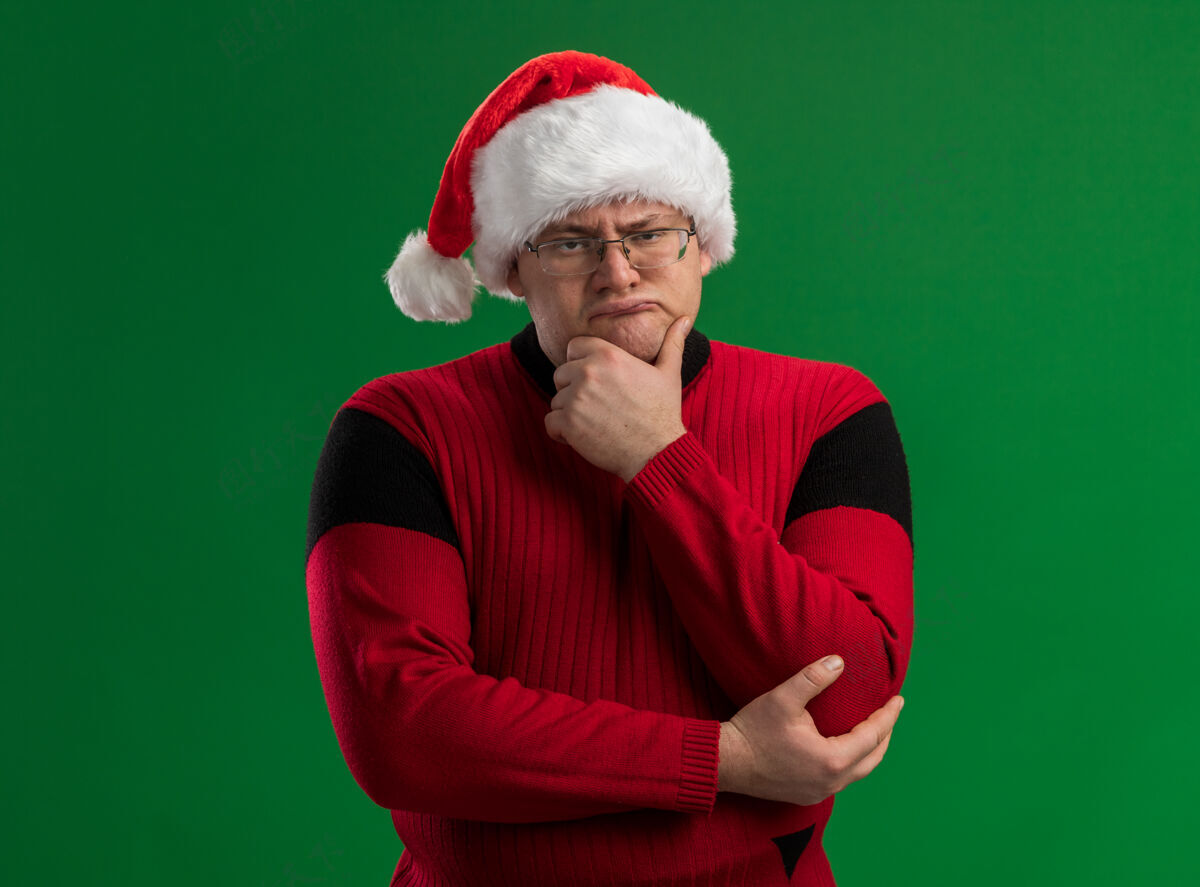 手皱眉的成年男子戴着眼镜 戴着圣诞老人的帽子 手放在下巴上 看着隔离在绿色背景下的相机皱眉眼镜成人