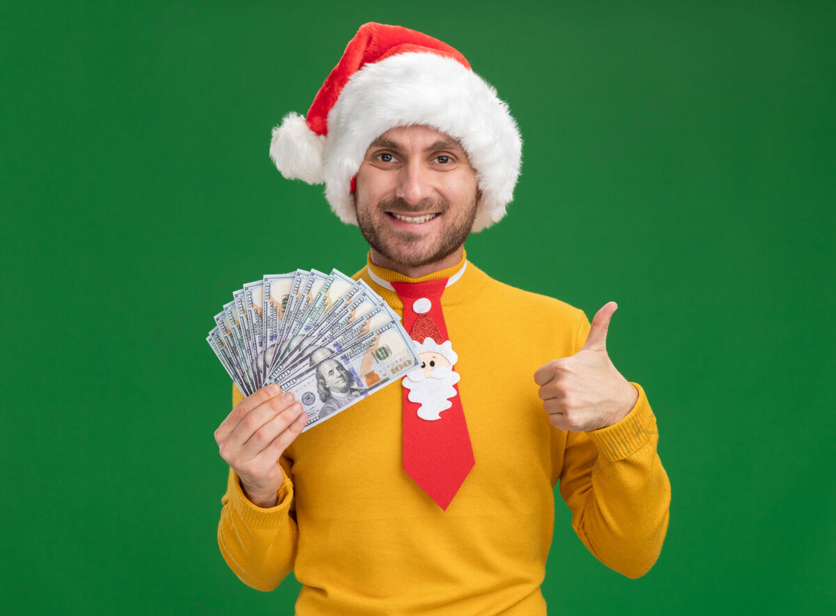 看微笑着的白人年轻人戴着圣诞帽 打着领带 手里拿着钱 看着相机 在绿色背景上孤立地竖起大拇指钱圣诞节年轻