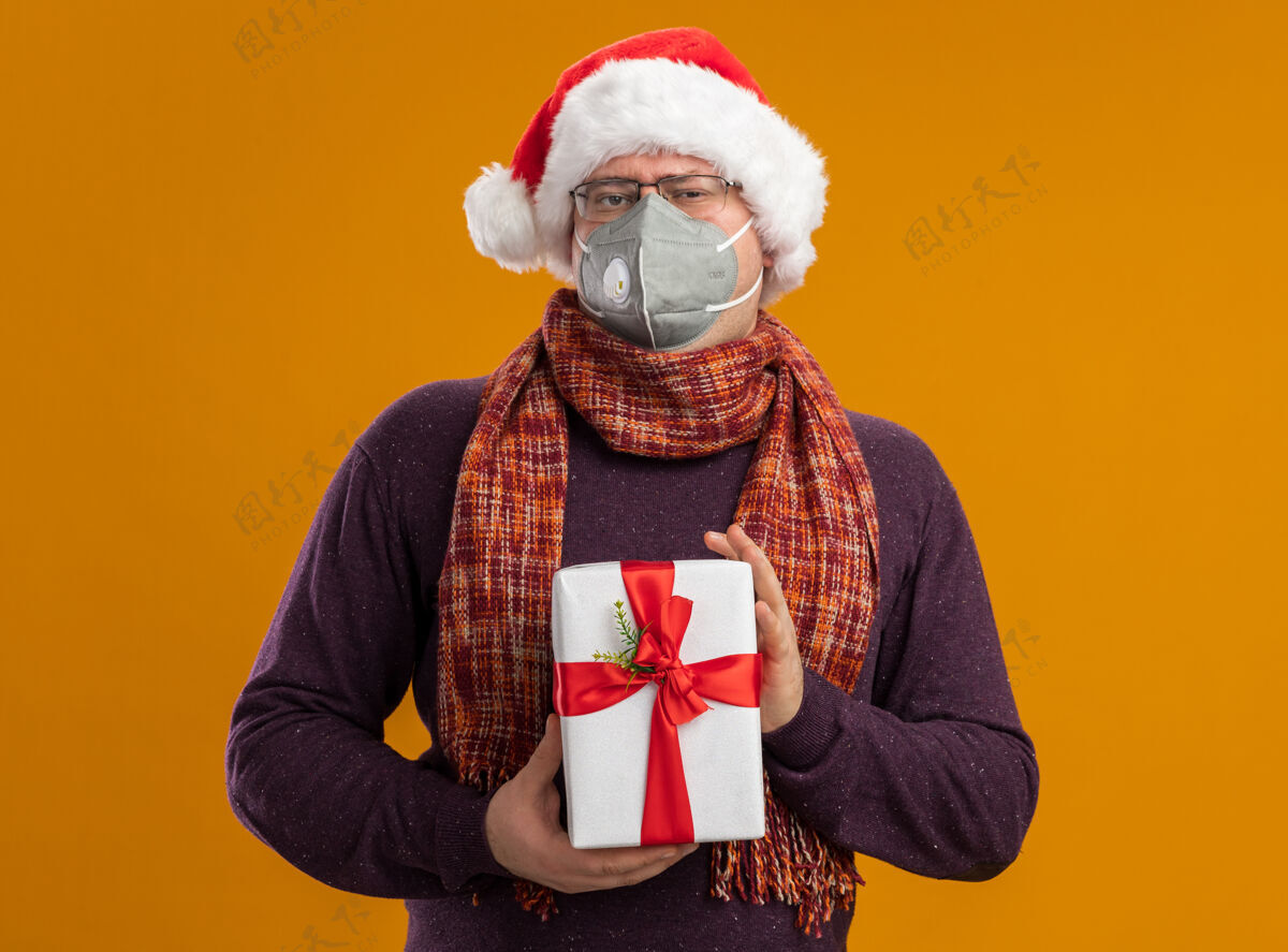 手捧自信的成年男子戴着防护面具 戴着眼镜 戴着圣诞帽 脖子上围着围巾 手里拿着礼包 看着隔离在橙色背景上的相机周围橙色帽子