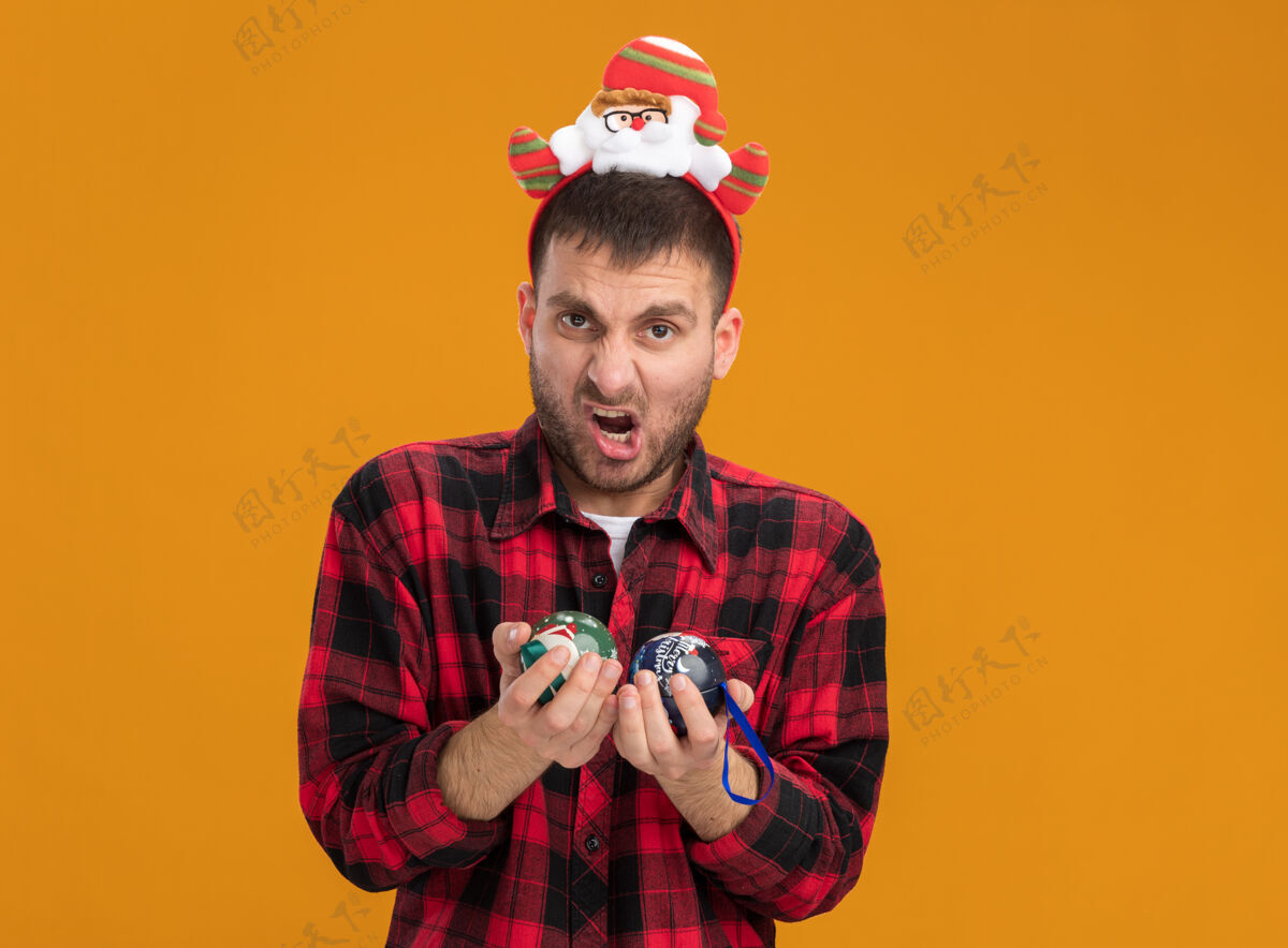 男人激进的白人年轻人戴着圣诞老人的头带 手里拿着圣诞饰品 隔离在橙色的墙上 留着复制空间穿着头带圣诞老人