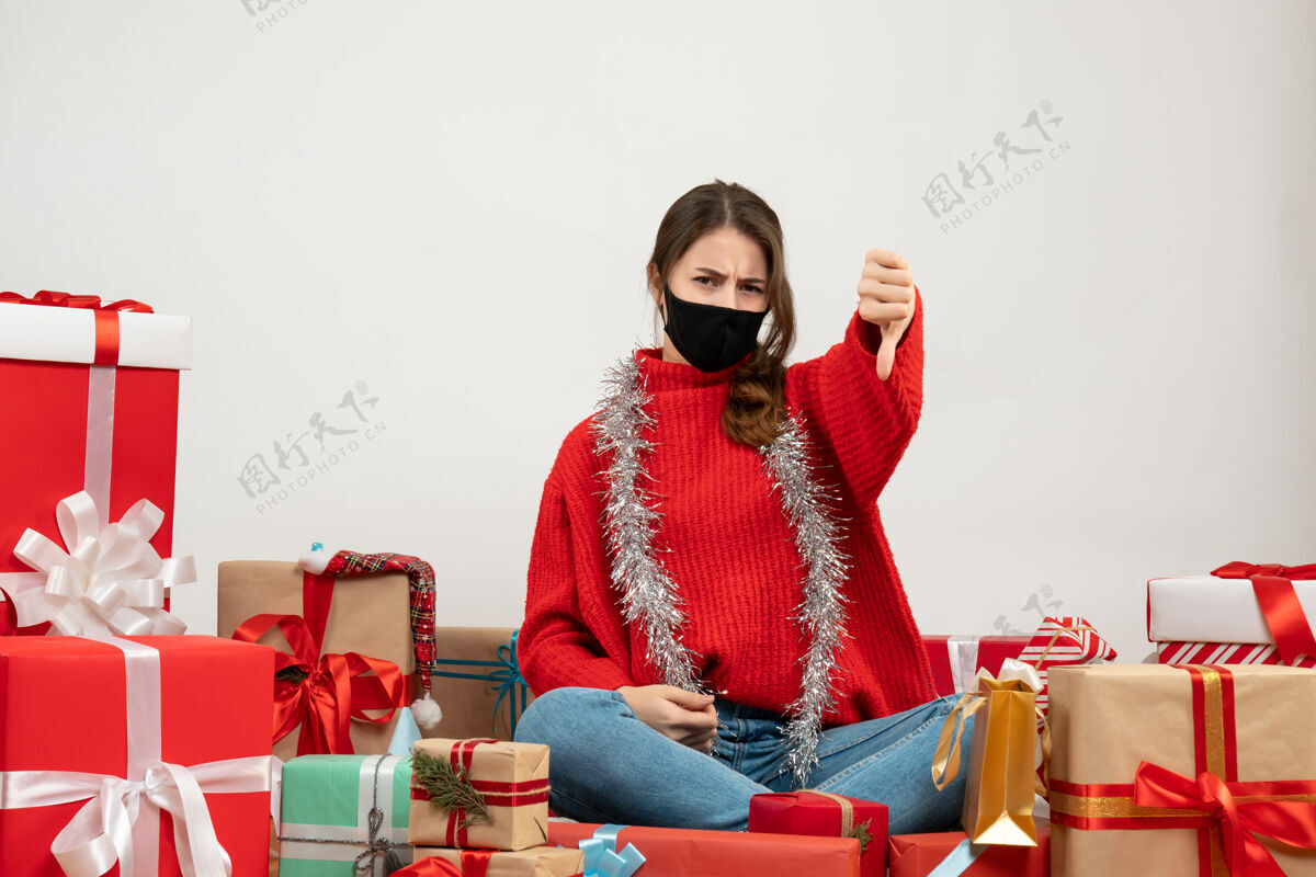 拇指穿着红色毛衣的年轻女孩做着拇指朝下的手势坐在礼物旁边 白色的面具是黑色的坐着肖像毛衣