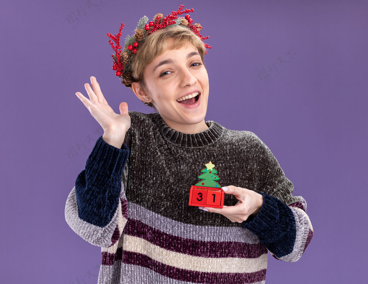 空虚快乐的年轻漂亮女孩戴着圣诞花环手持圣诞树玩具和日期显示空手孤立在紫色的墙上头花环美丽