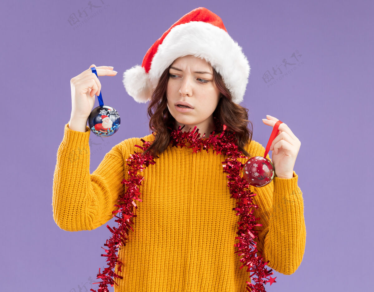 脖子无知的年轻斯拉夫女孩 戴着圣诞帽 脖子上戴着花环 拿着玻璃球 看着紫色背景上的装饰物 还有复制空间周围斯拉夫圣诞老人