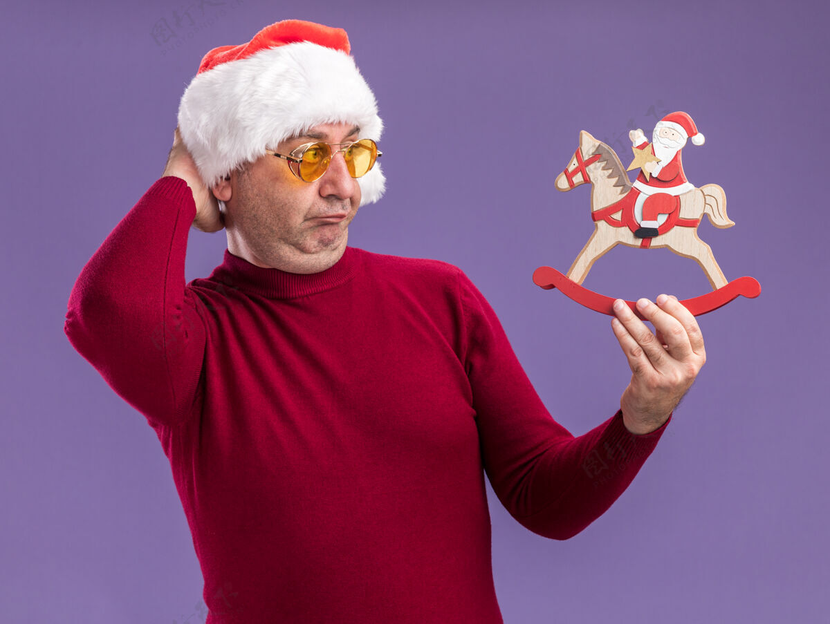 中间中年男子戴着圣诞老人帽 戴着黄色眼镜 手里拿着圣诞玩具 困惑地看着它 手放在头上 站在紫色的背景上帽子头手
