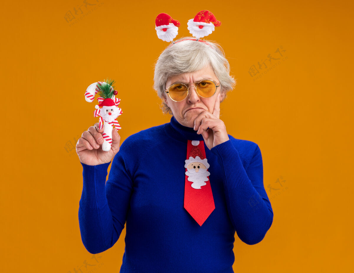 眼镜戴着太阳眼镜 戴着圣诞老人头带和圣诞老人领带的恼怒的老妇人把手放在下巴上 拿着橙色背景上的糖果手杖 留有复印空间烦恼橘子糖果