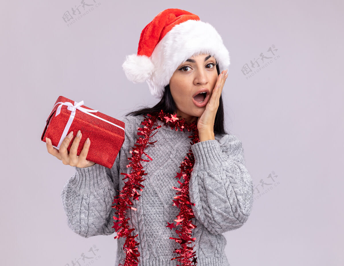 圣诞节关心的年轻白人女孩戴着圣诞帽 脖子上戴着金箔花环 手里拿着礼包 手放在脸上 隔离在白色的墙上 留着复印空间包装周围保管