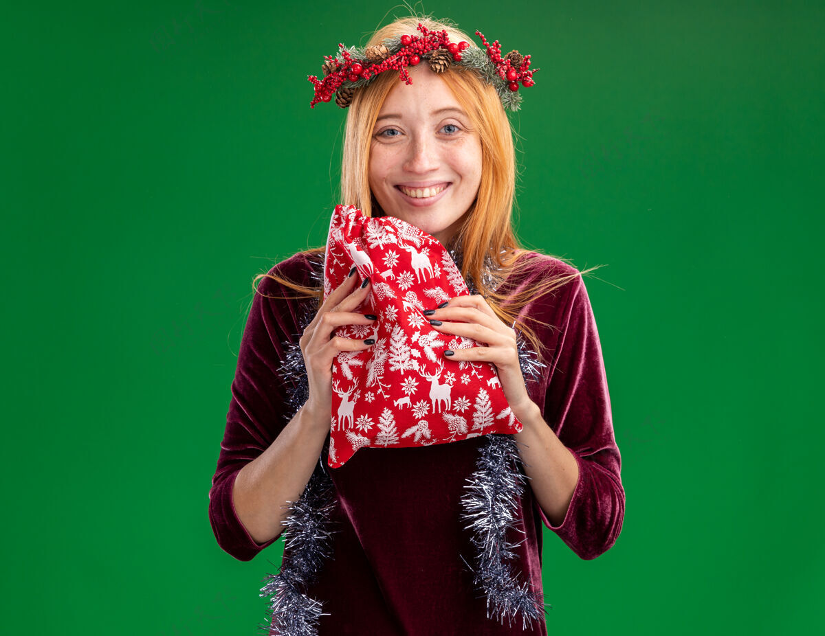 女孩微笑着的年轻漂亮的女孩穿着红色的裙子 脖子上戴着花环和花环 手里拿着绿色背景上孤立的圣诞包花环微笑衣服