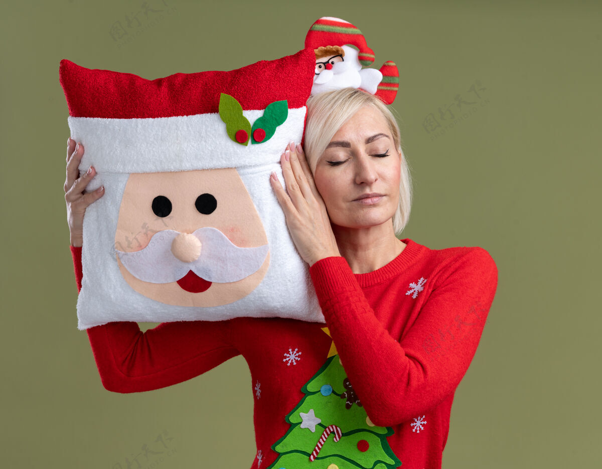 枕头放松的中年金发女人戴着圣诞老人的头带和圣诞毛衣抱着圣诞老人的枕头摸着它的头 闭着眼睛隔离在橄榄绿的背景上圣诞老人眼睛橄榄
