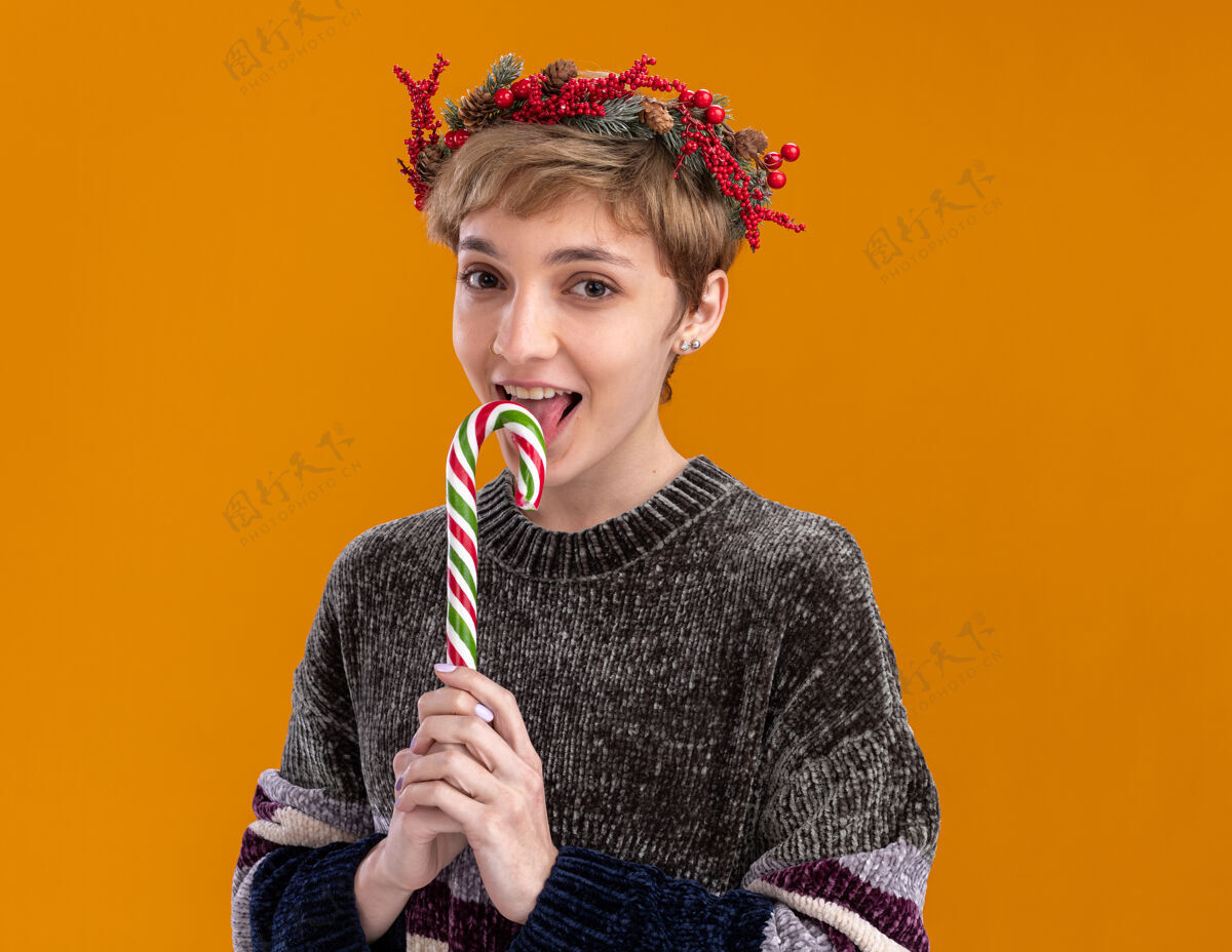 头快乐的年轻漂亮女孩戴着圣诞花环拿着圣诞甜甜的手杖舔着隔离在橙色墙上的复制空间快乐花环年轻