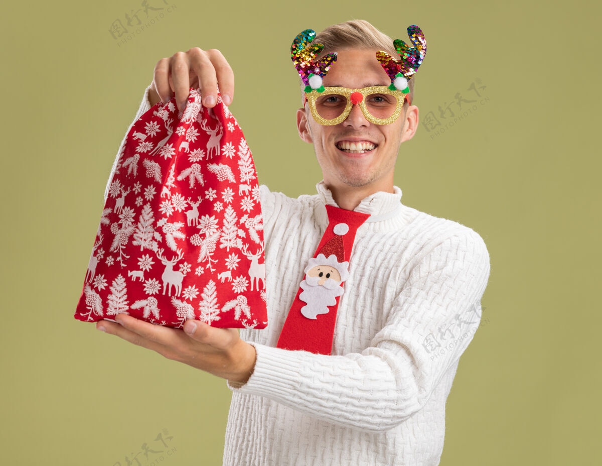 年轻微笑着的年轻帅哥戴着圣诞新奇眼镜 打着圣诞老人的领带 把圣诞袋伸向相机 看着橄榄绿背景上孤立的相机小伙子微笑看