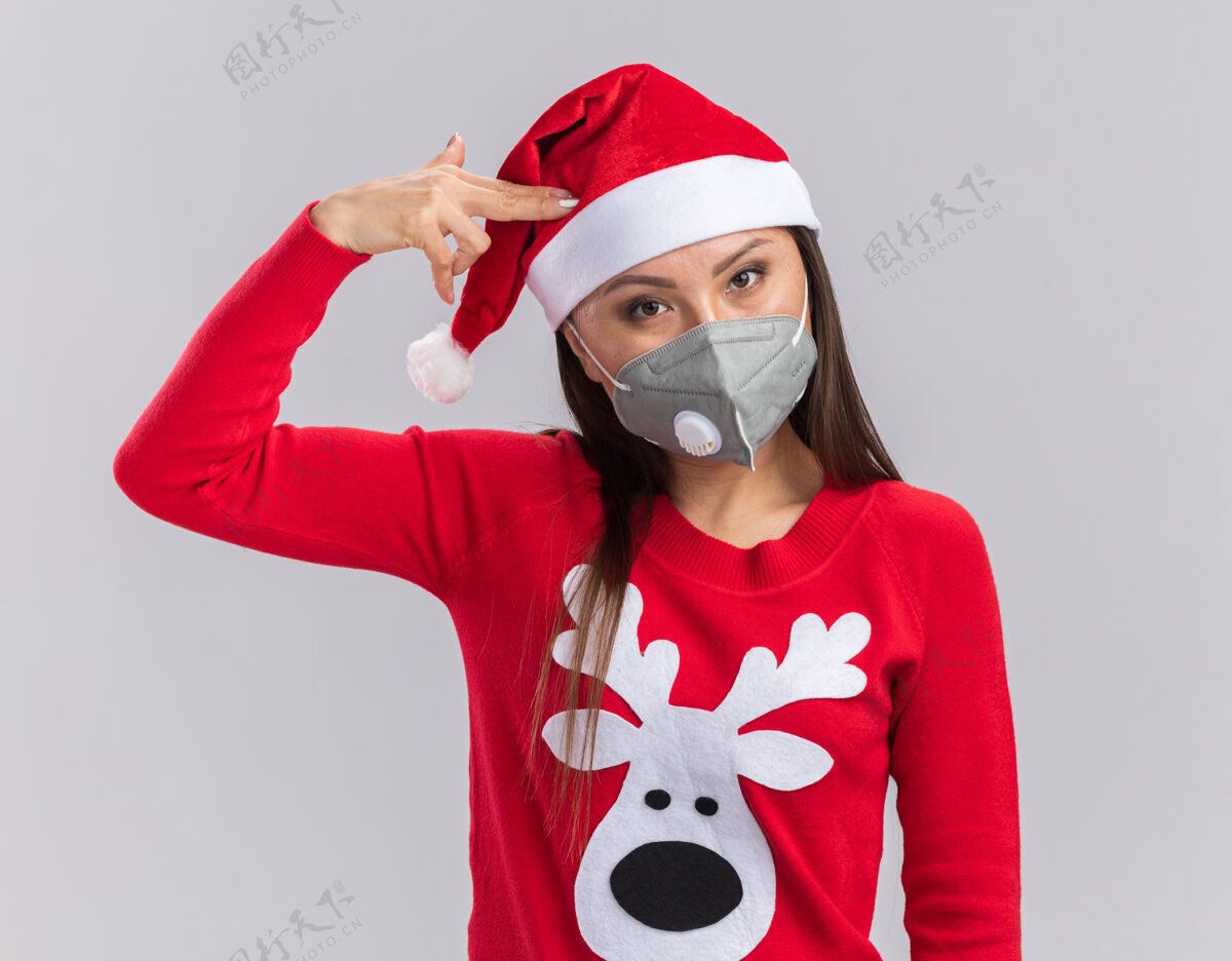 年轻看着镜头 年轻的亚洲女孩戴着圣诞帽 穿着毛衣 戴着医用面罩 在白色的背景上显示出的姿势亚洲人医疗帽子