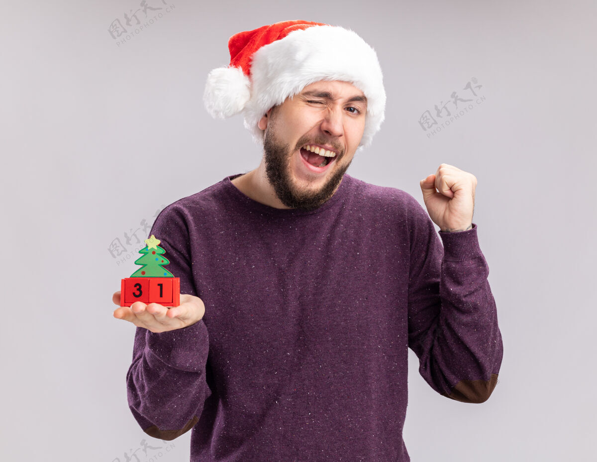 毛衣身穿紫色毛衣 头戴圣诞帽的年轻人站在白色背景上 展示着握紧新年拳头的方块帽子站着立方体