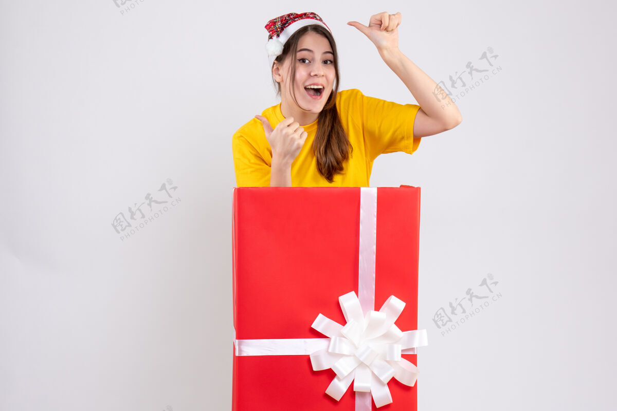 拇指圣诞快乐女孩戴着圣诞帽 竖起大拇指 站在白色圣诞礼物后面大帽子圣诞节