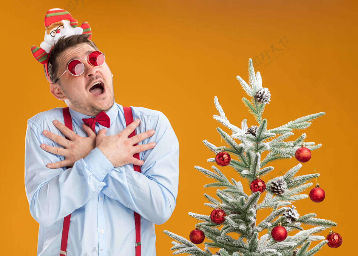 胸部戴着吊带领结的年轻人站在圣诞树旁 戴着圣诞老人和红眼镜 在橙色的背景下 手放在胸前 惊讶而担忧地抬头望着吊带手圣诞老人
