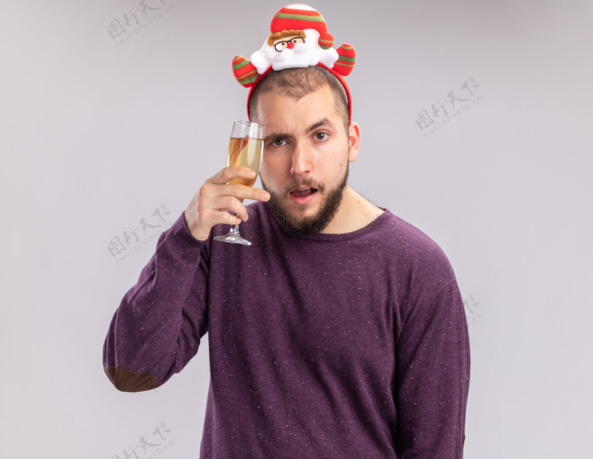 站着快乐的年轻人穿着紫色毛衣 头上戴着滑稽的边 手里拿着一杯香槟 站在白色的背景下 看上去很自信年轻香槟毛衣