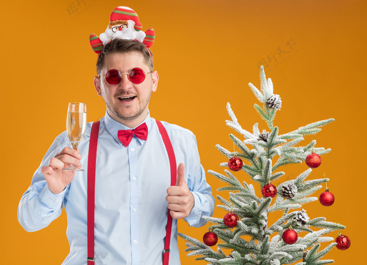 香槟戴吊带领结的年轻人站在圣诞树旁 戴着圣诞老人和红眼镜 手里拿着香槟酒杯 竖起大拇指 在橙色的背景下快乐快乐吊带玻璃领带