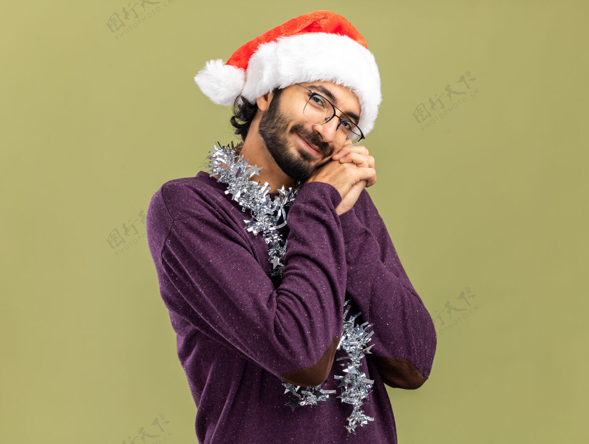 帅气高兴的年轻帅哥戴着圣诞帽 脖子上戴着花环 手放在脸颊上 隔离在橄榄绿的背景上橄榄抱着高兴