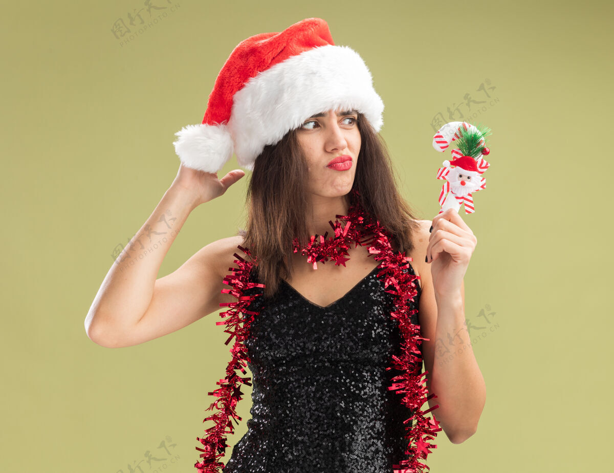 年轻不愉快地看着身边年轻漂亮的女孩戴着圣诞帽 脖子上戴着花环 手里拿着橄榄绿背景上孤立的圣诞玩具颈部未发行举行