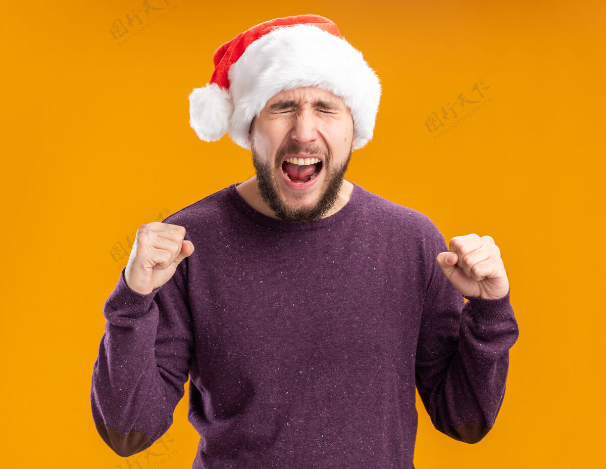 兴奋穿着紫色毛衣 戴着圣诞帽的年轻人站在橙色的背景上 兴奋地握紧拳头 高喊着疯狂的快乐握紧帽子站着