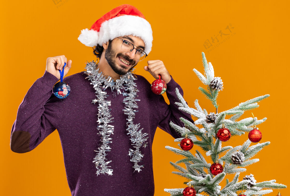 倾斜微笑着歪着头的年轻帅哥站在圣诞树旁 戴着圣诞帽 脖子上戴着花环 手里拿着橙色背景上孤立的圣诞球脖子头帅气