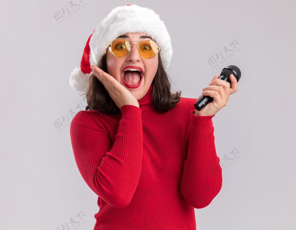 圣诞老人一个穿着红毛衣 戴着圣诞帽 戴着眼镜 拿着麦克风 站在白色背景上看镜头的快乐而兴奋的小女孩站着眼镜兴奋