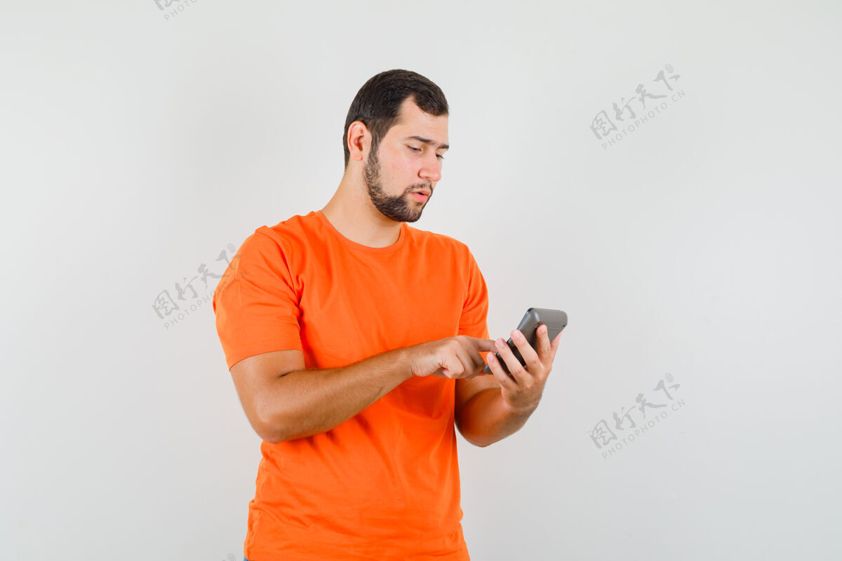 休闲年轻人正在计算器上做计算 穿着橙色t恤 看上去很忙 正面视图成人男人时尚