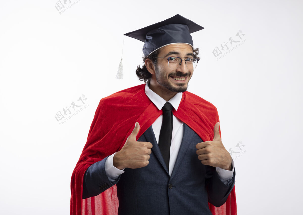 复制微笑着的年轻白种人超级英雄 戴着眼镜 穿着红色斗篷的西装 戴着毕业帽 竖起大拇指 两只手孤立地放在白色背景上 留着复印空间年轻毕业帽子