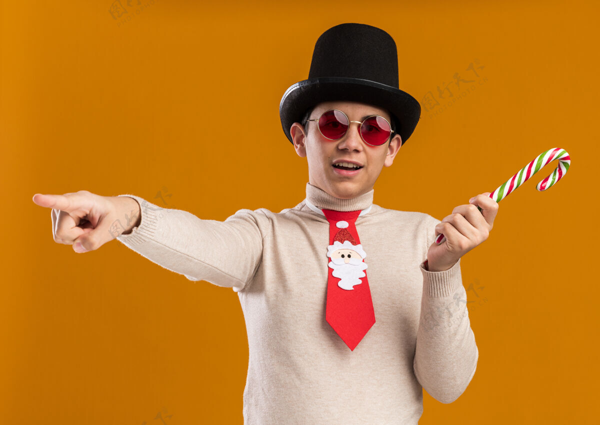 糖果高兴的年轻人戴着帽子 戴着圣诞领带 戴着一副眼镜 手里拿着圣诞糖果点 隔离在黄色背景上领带点举行