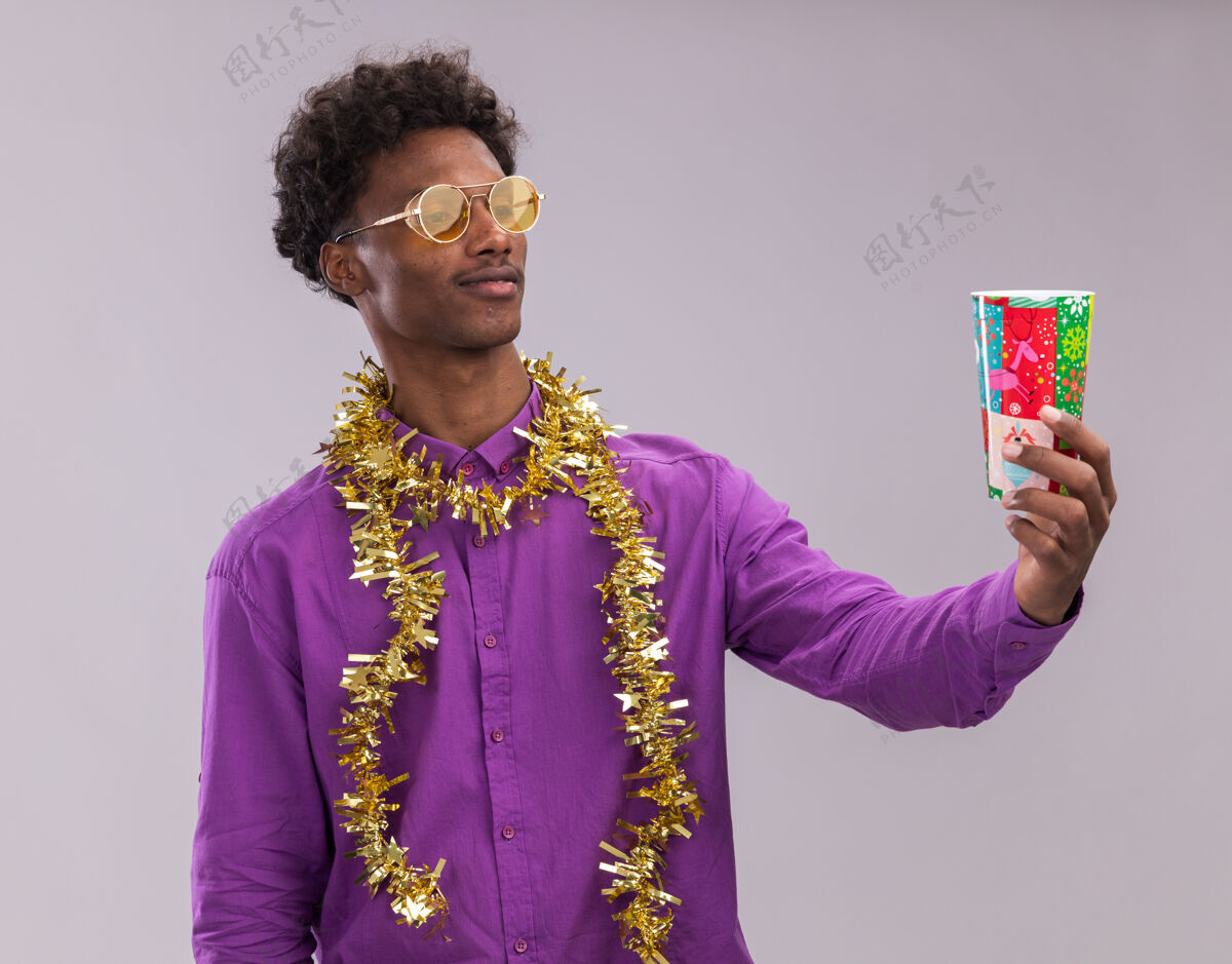 年轻困惑的美国黑人年轻人戴着眼镜 脖子上戴着金属丝花环 伸出塑料圣诞杯 在白色背景上孤立地看着它杯子周围圣诞节