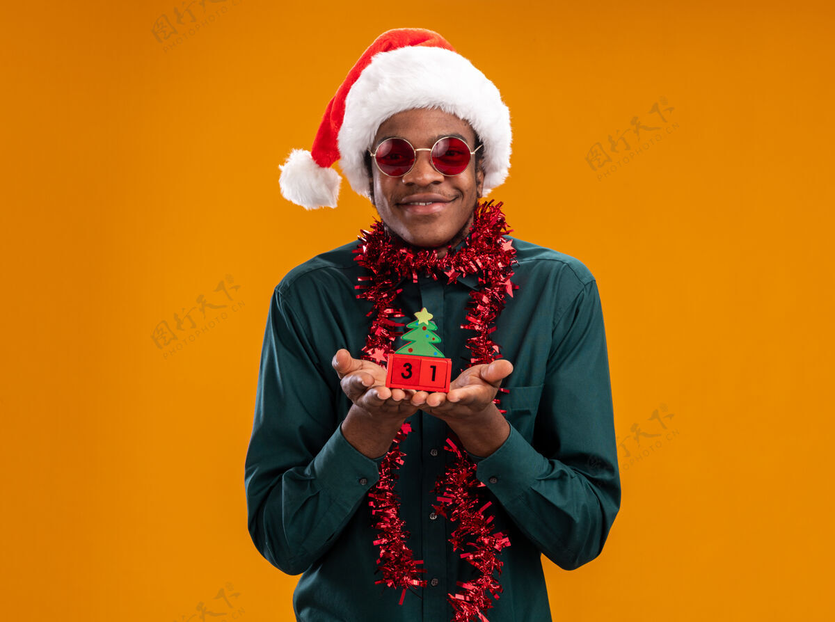 花环快乐的非洲裔美国人戴着圣诞帽 戴着花环 戴着太阳镜 抱着新年约会的玩具立方体 站在橙色背景下 看着相机 开心地微笑着立方体高兴美国