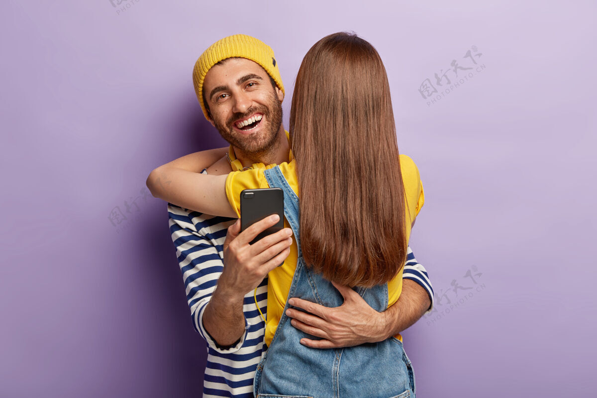 手机快乐的男性年轻人在网上聊天的时间与少女 拥抱妇女谁站在背后的镜头 享受生活 检查信息的追随者媒体在一起手机
