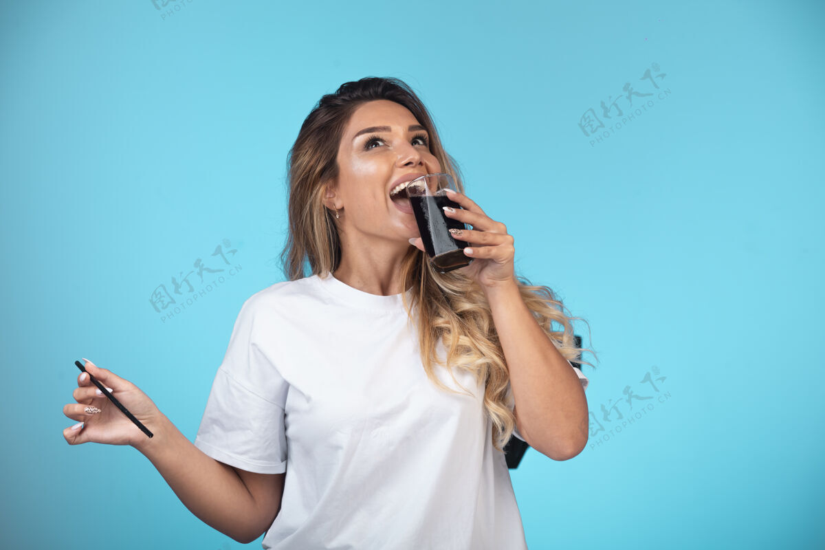 休闲穿白衬衫的年轻女孩喝着一杯黑鸡尾酒摆姿势年轻人茶点