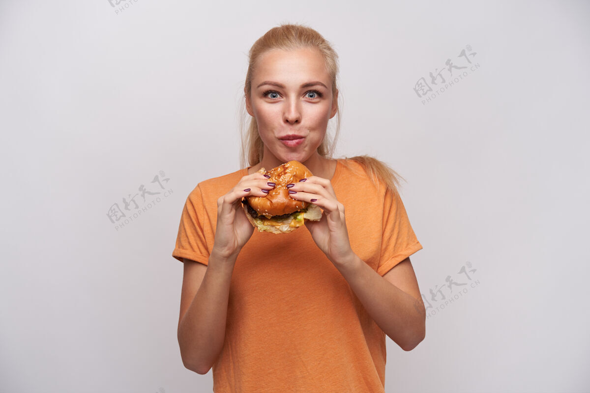 食欲一张室内照片 迷人的饥饿金发女郎 扎着马尾辫 嚼着美味的汉堡包 兴奋地看着镜头 身着白色背景的橙色t恤汉堡汉堡好