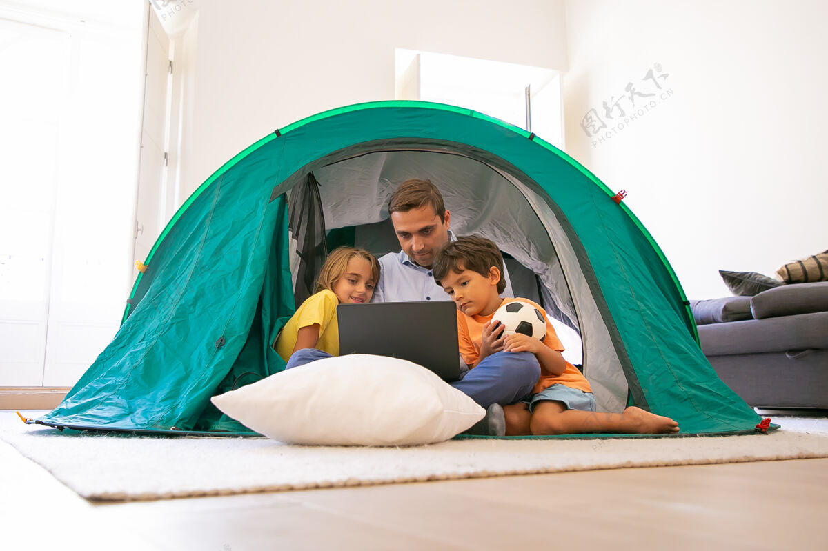 女儿有爱心的爸爸和孩子们盘腿坐在家里的帐篷里 看着笔记本电脑屏幕可爱的孩子们和爸爸一起在电脑上看电影童年 家庭时间和周末的概念男孩关心盘腿