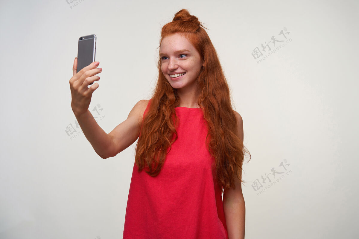 制作一个积极的年轻readhead女士的工作室照片 她卷曲的红色头发在白色背景下摆姿势 不化妆 穿着休闲的粉色连衣裙在智能手机上自拍室内发髻牙齿