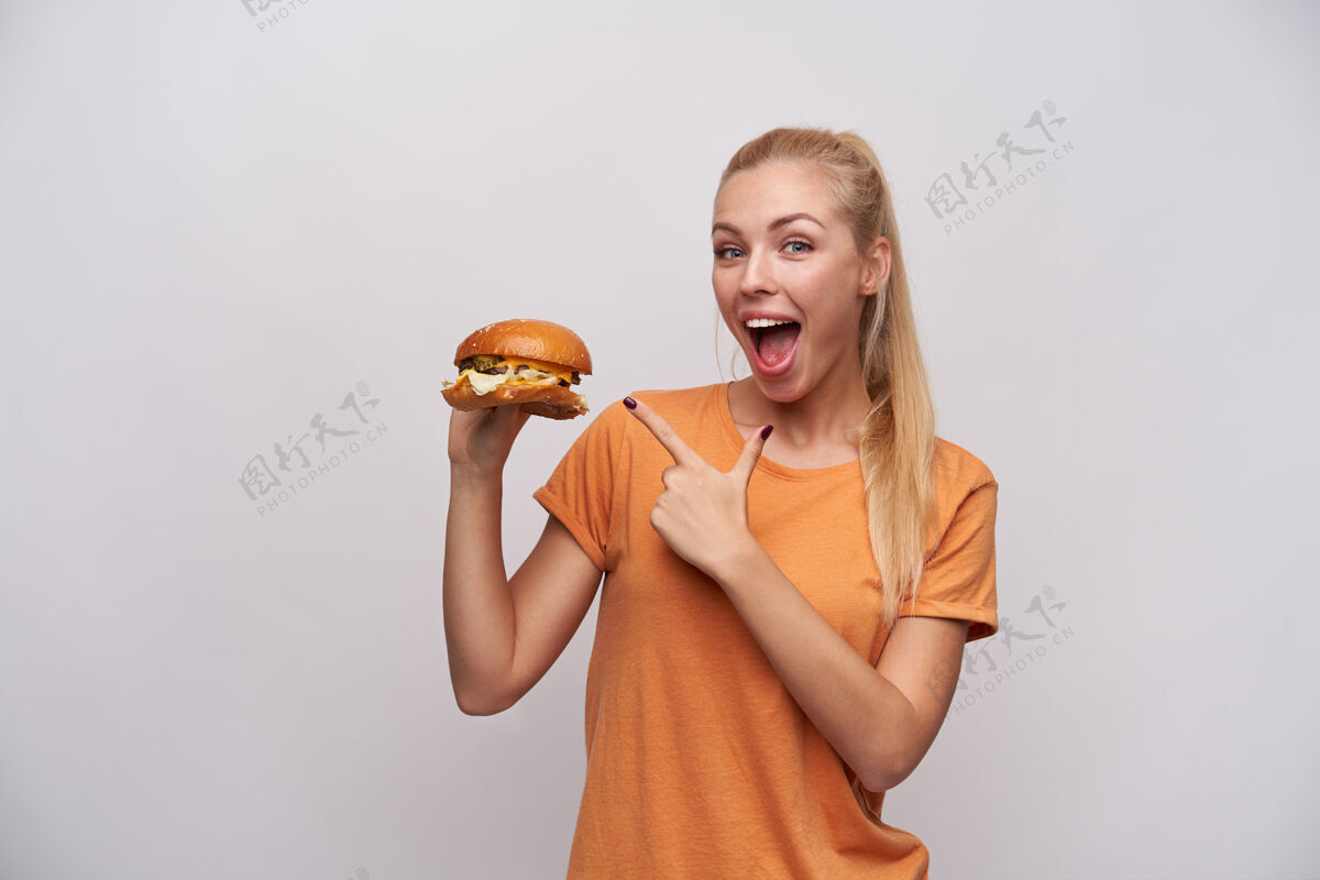 20多岁一位穿着休闲服的年轻金发女郎兴奋地抬起手 用食指指着新鲜的大汉堡 在白色背景下摆出一副开心的姿势 面带微笑地看着镜头金发漂亮表情