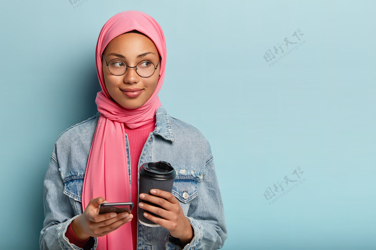 短信科技和通讯理念身着粉色面纱的穆斯林妇女的照片 使用新安装的智能手机应用程序 端着咖啡走 戴着圆眼镜 站在室内蓝色墙壁上宗教个性业余时间