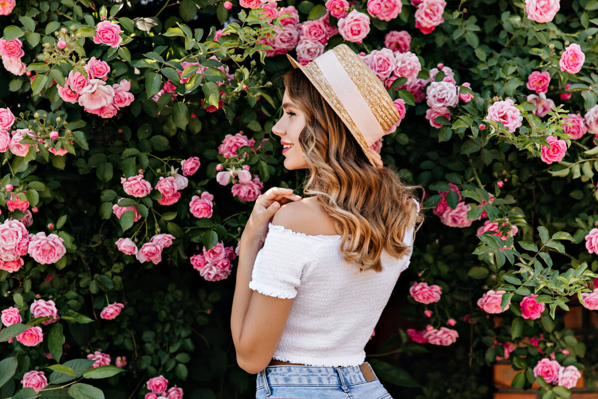 自然美丽的高加索女孩看着粉色玫瑰的照片戴帽子的时尚女模特站在花丛旁的户外照片草帽花微笑